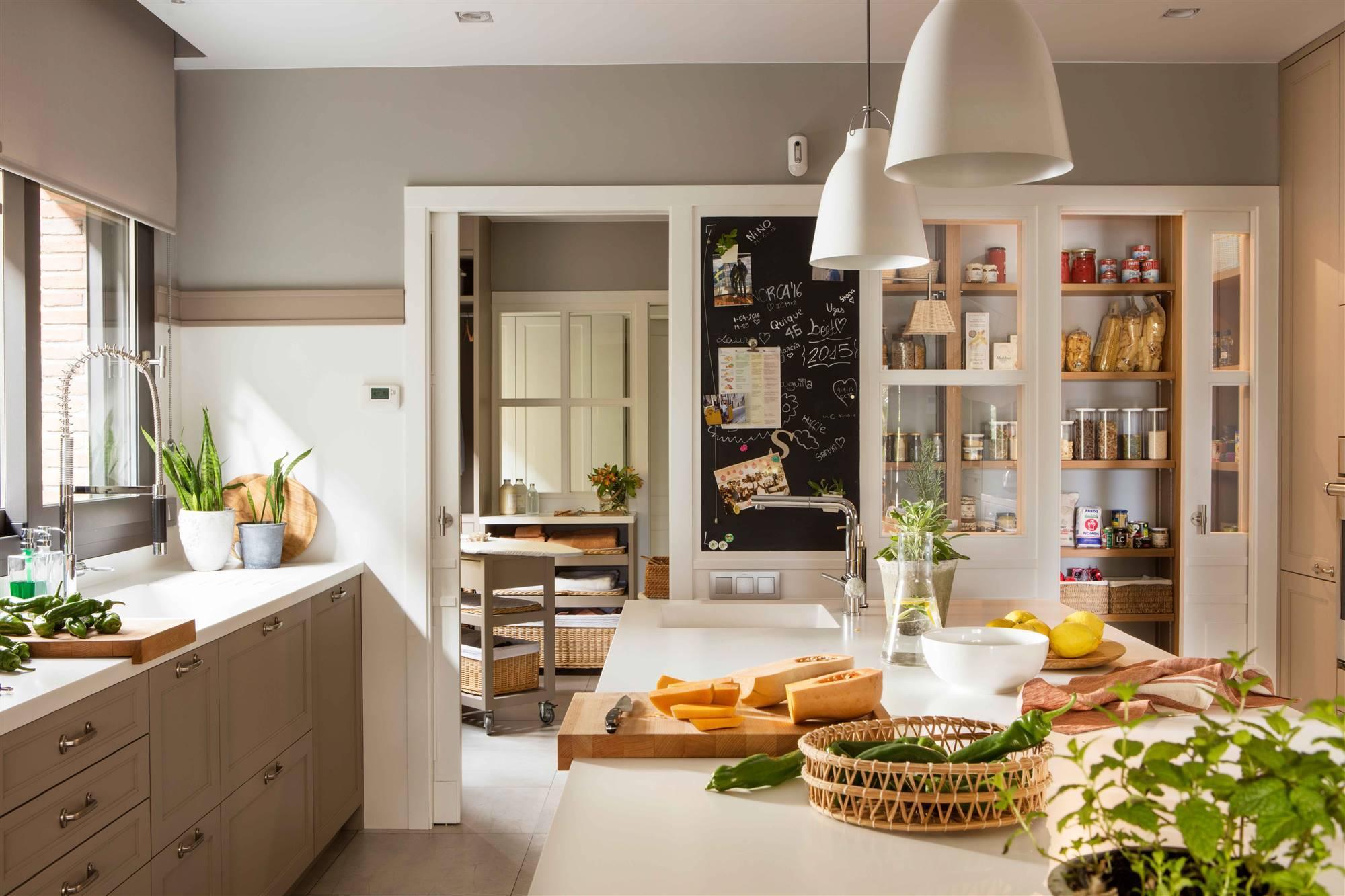 A bright, modern kitchen interior with light gray cabinets, white countertops, pendant lights, glass-front pantry shelves, and a central island with food items and plants.